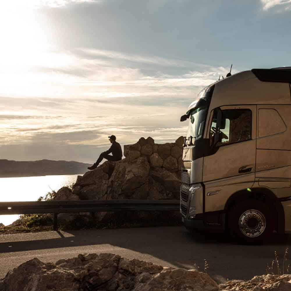 En chauffør sidder på en klippe og ser ud over havet ved solnedgang med sin Volvo FH-lastbil parkeret ved siden af